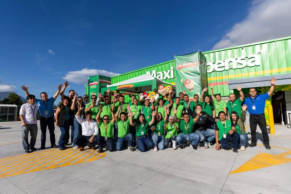 Gran equipo de empleados sonrientes en uniformes verdes posa frente a una tienda 'Maxi Pensa' con carteles de '¡Ya abrimos!' y 'Jocotenango', celebrando una apertura.