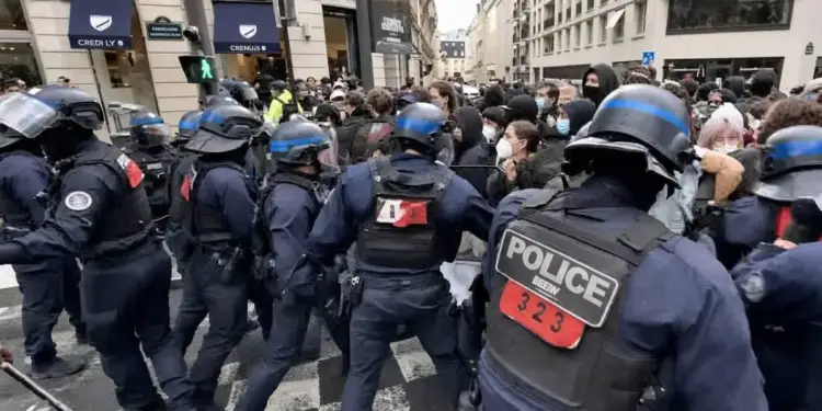Agentes de policía con equipo antidisturbios y cascos se encuentran frente a una multitud de personas en una calle urbana con tiendas al fondo.