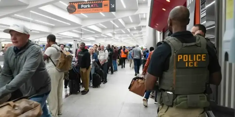 Vista trasera de un agente de ICE con chaleco táctico en un aeropuerto concurrido, donde pasajeros con equipaje esperan en fila. Se ve un cartel de "Park ATL".