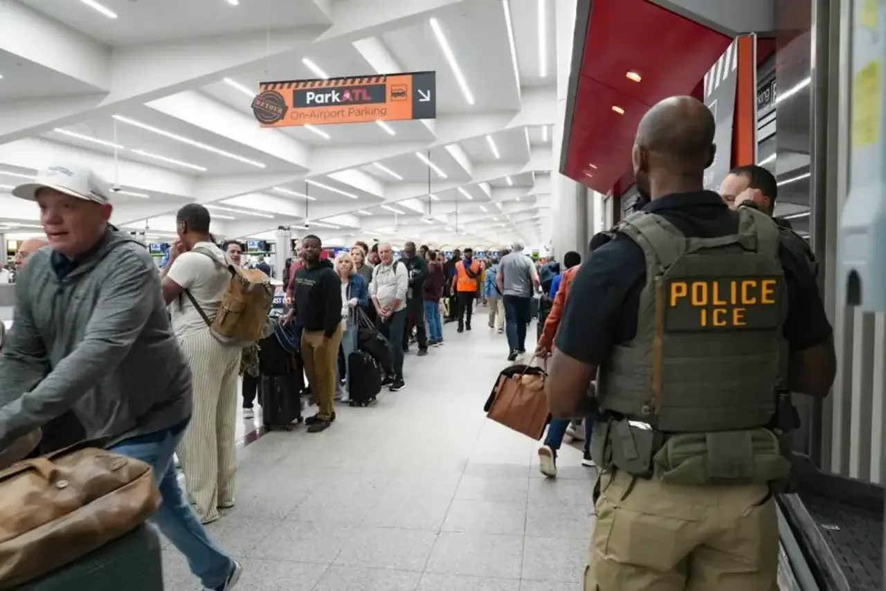 Vista trasera de un agente de ICE con chaleco táctico en un aeropuerto concurrido, donde pasajeros con equipaje esperan en fila. Se ve un cartel de "Park ATL".