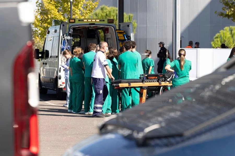 Grupo de profesionales de la salud con uniformes verdes y un paramédico junto a una ambulancia con puertas abiertas y una camilla. Escena de emergencia médica al aire libre.