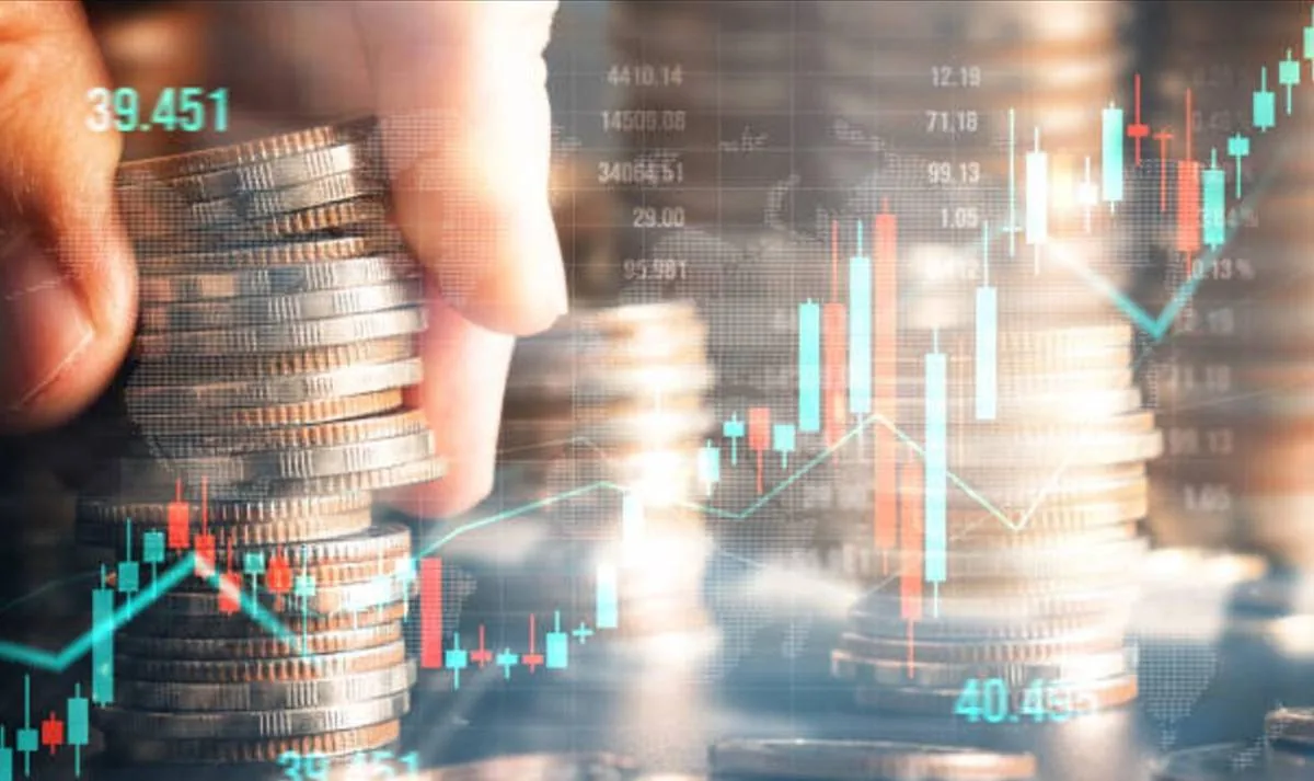 Mano apilando monedas de diferentes metales sobre un fondo de gráficos bursátiles y datos financieros digitales, simbolizando inversión y crecimiento económico.