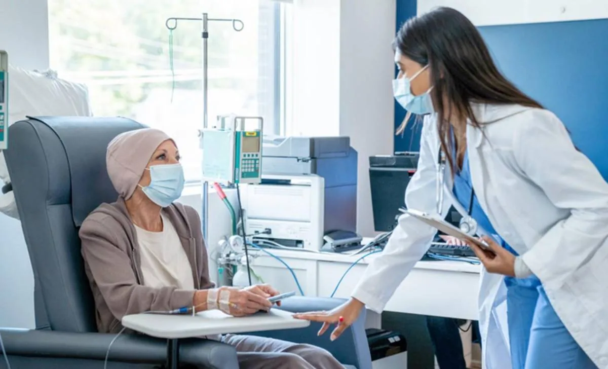 Médica con mascarilla conversa con una paciente en sillón reclinable recibiendo tratamiento médico.