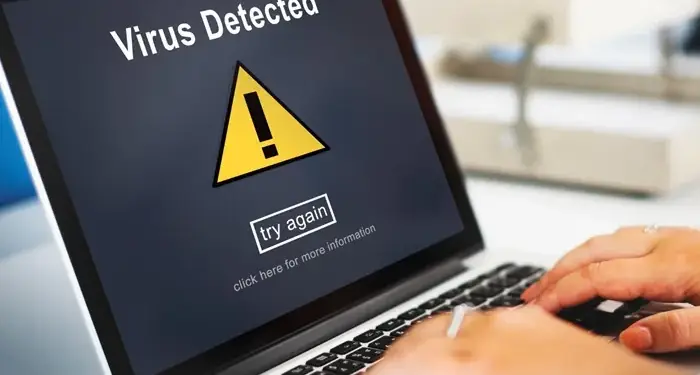 Manos escribiendo en un ordenador portátil que muestra una alerta de virus detectado con un símbolo de advertencia.