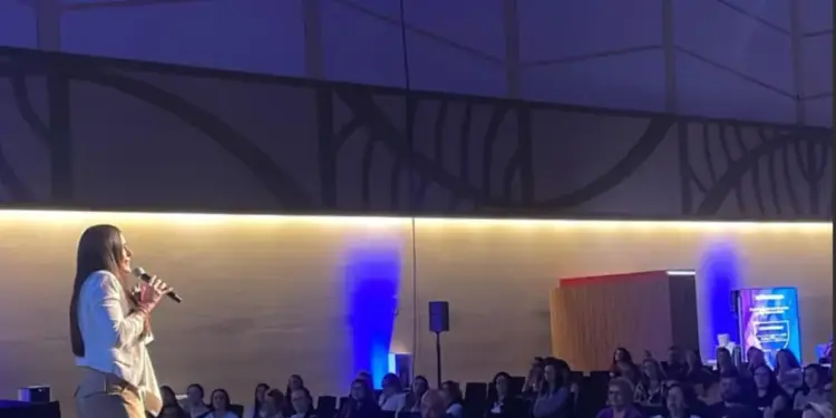 Una mujer con un traje blanco y pantalones beige habla en un micrófono frente a una audiencia sentada en un auditorio con luces azules.