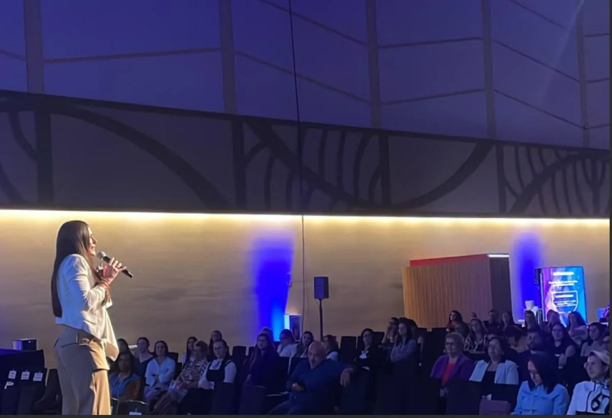 Una mujer con un traje blanco y pantalones beige habla en un micrófono frente a una audiencia sentada en un auditorio con luces azules.