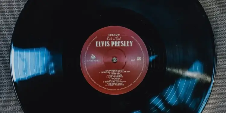 Primer plano de un disco de vinilo negro de Elvis Presley con etiqueta roja, mostrando títulos de canciones y detalles de derechos de autor. Reflejos azules en la superficie.