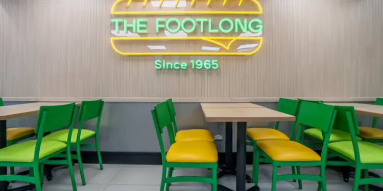 Interior de restaurante con mesas de madera y sillas verdes/amarillas. Letrero de neón "THE FOOTLONG Since 1965" en pared de paneles.