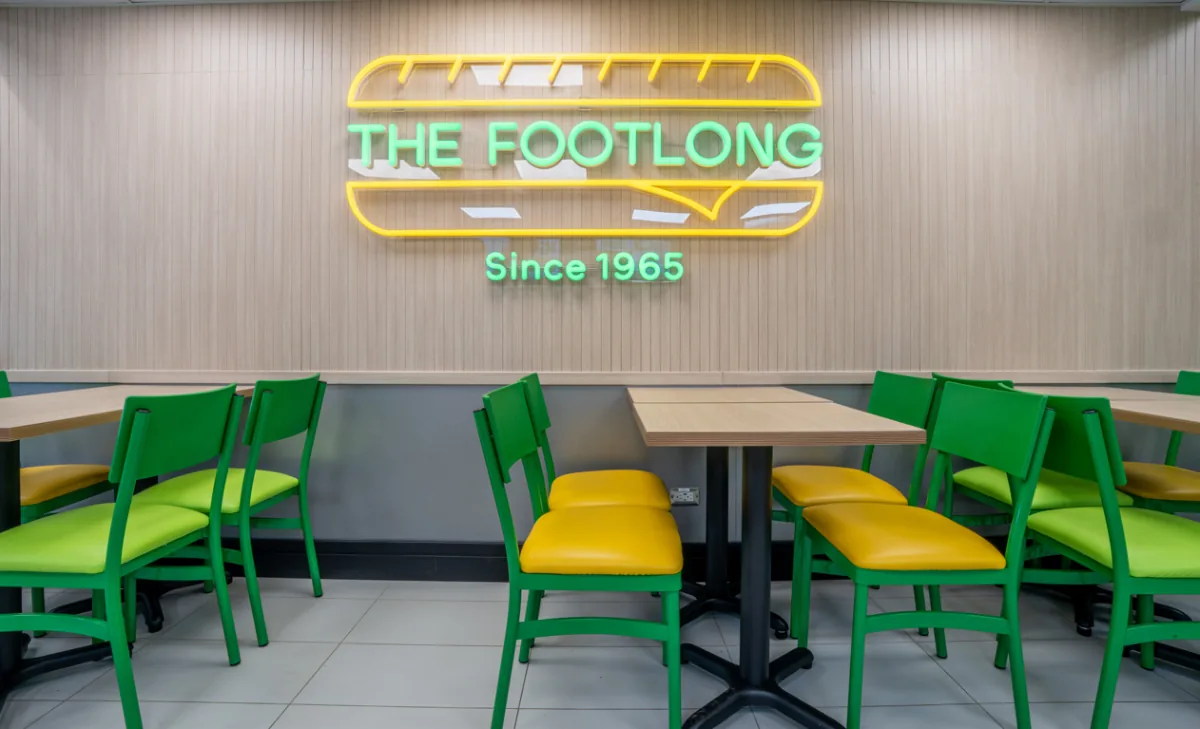 Interior de restaurante con mesas de madera y sillas verdes/amarillas. Letrero de neón "THE FOOTLONG Since 1965" en pared de paneles.