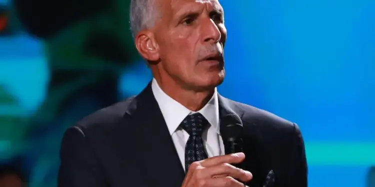 Hombre mayor en traje oscuro y corbata sosteniendo un micrófono mientras habla en una conferencia con fondo azul.