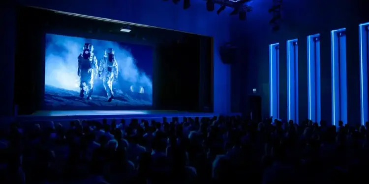Una audiencia observa una pantalla grande que muestra a dos astronautas caminando en un paisaje lunar, en una sala con iluminación azul.
