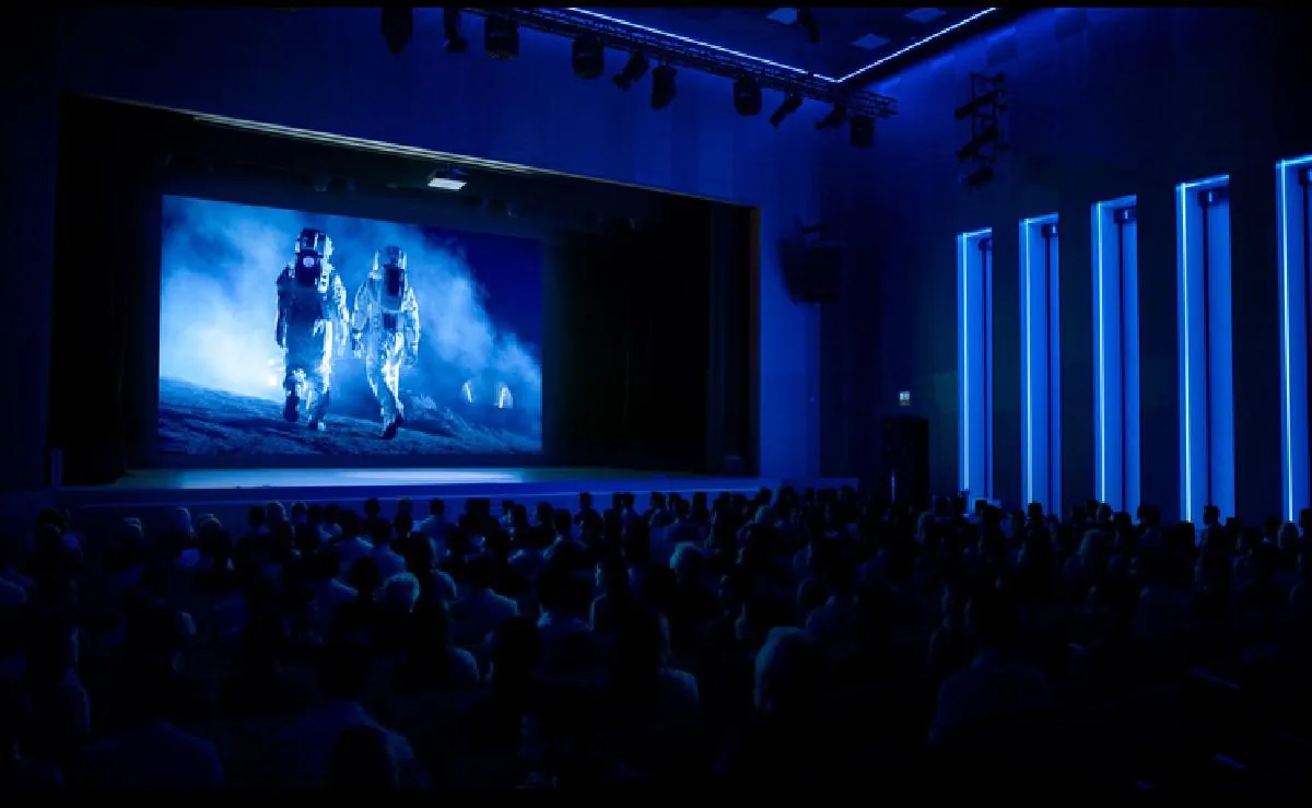 Una audiencia observa una pantalla grande que muestra a dos astronautas caminando en un paisaje lunar, en una sala con iluminación azul.