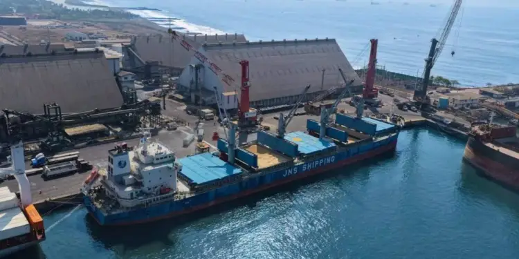 Vista aérea de un gran buque carguero atracado en un puerto industrial con grúas y grandes almacenes junto al mar.