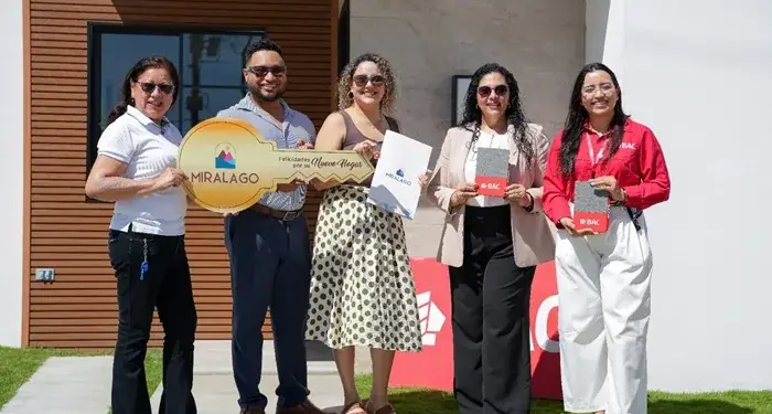 Cinco personas, tres mujeres y dos hombres, celebran la entrega de una vivienda. Sostienen una llave gigante de 'MIRALAGO' y documentos de 'BAC'.