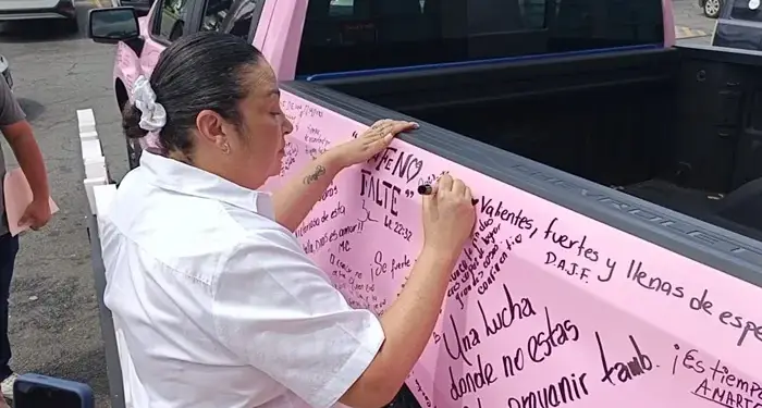 Mujer con camisa blanca escribiendo mensajes motivacionales con marcador en la superficie lateral de una camioneta rosa.