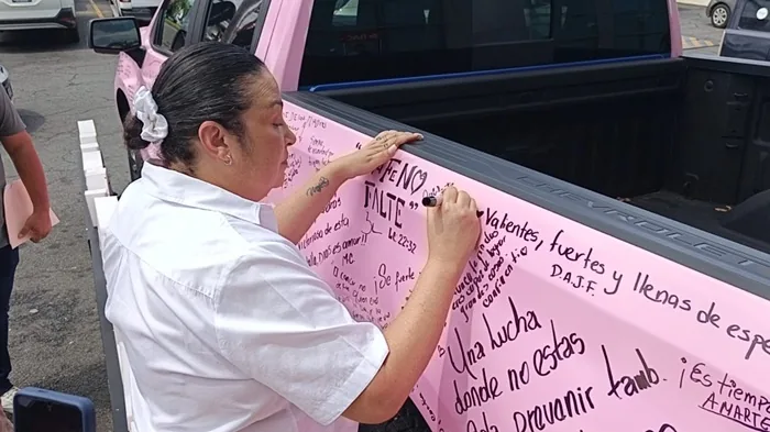 Mujer con camisa blanca escribiendo mensajes motivacionales con marcador en la superficie lateral de una camioneta rosa.