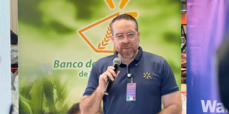 Hombre con barba y gafas hablando por un micrófono frente a un cartel corporativo.