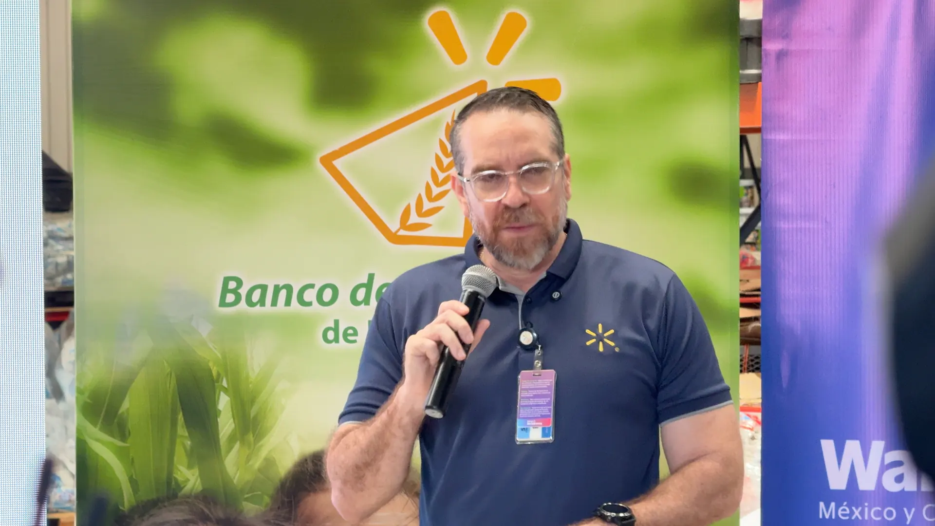 Hombre con barba y gafas hablando por un micrófono frente a un cartel corporativo.