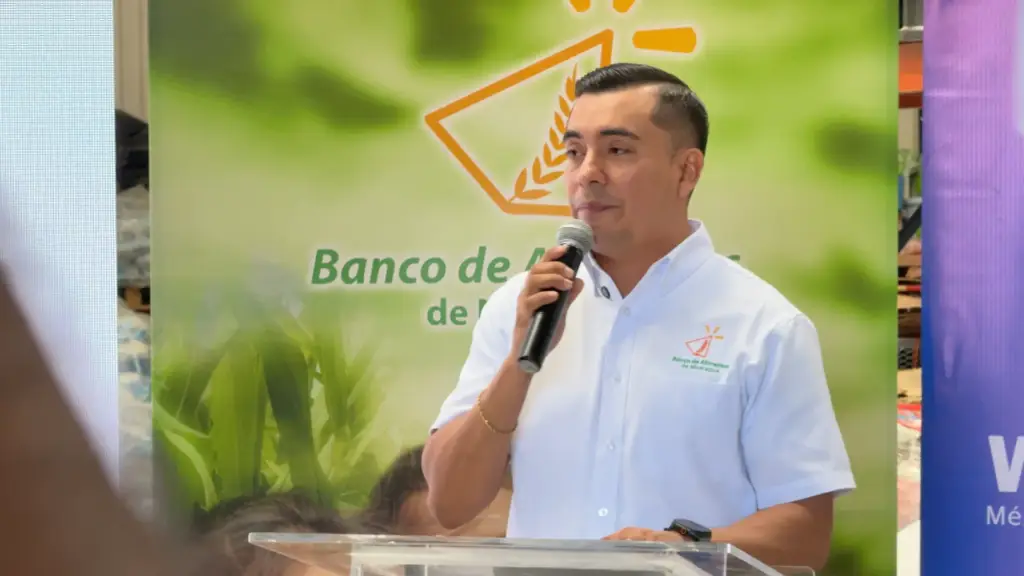 Hombre con camisa blanca hablando frente a un atril con un micrófono en la mano y un cartel verde de fondo.