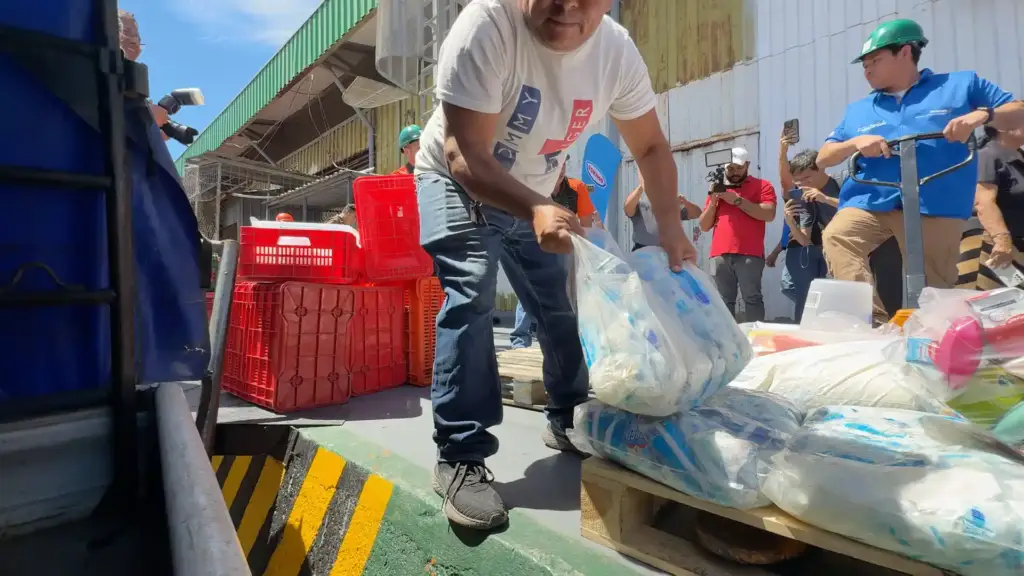 Hombre descargando bolsas de productos en una zona de carga junto a estibas con mercancía y trabajadores.