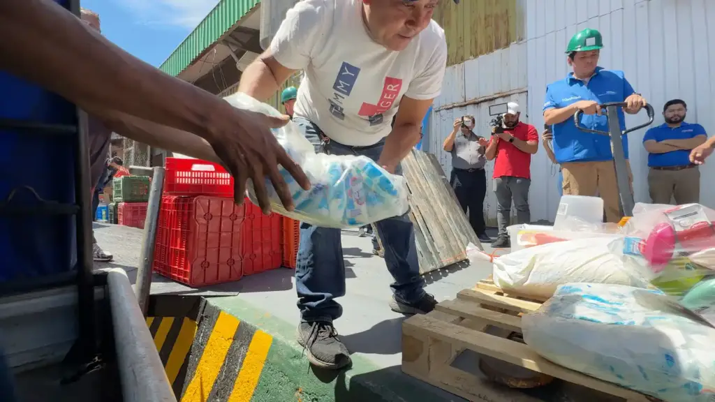 Hombre entregando una bolsa con productos a otra persona en un área de carga con tarimas de madera y cajas rojas.