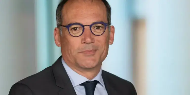 Retrato de un hombre de mediana edad con traje oscuro, camisa azul claro y corbata oscura. Lleva gafas de montura azul y tiene una expresión seria. Fondo borroso de oficina.