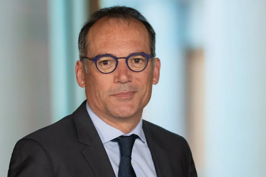 Retrato de un hombre de mediana edad con traje oscuro, camisa azul claro y corbata oscura. Lleva gafas de montura azul y tiene una expresión seria. Fondo borroso de oficina.