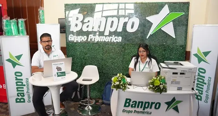 Un hombre y una mujer sentados en un stand con computadoras portátiles frente a un muro verde con el logotipo de Banpro Grupo Promerica.