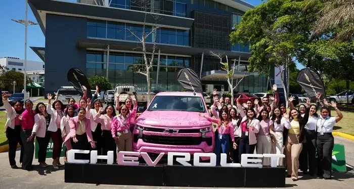 Grupo de personas sonriendo y levantando las manos frente a una camioneta rosa y un edificio corporativo moderno.