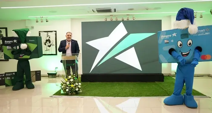 Hombre en podio con micrófono, flanqueado por dos mascotas de tarjetas de crédito Banpro en un evento corporativo con logo de estrella.
