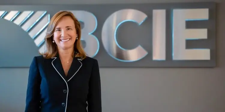 Mujer profesional sonriente vestida con traje formal oscuro frente al logotipo metálico de BCIE en una oficina.