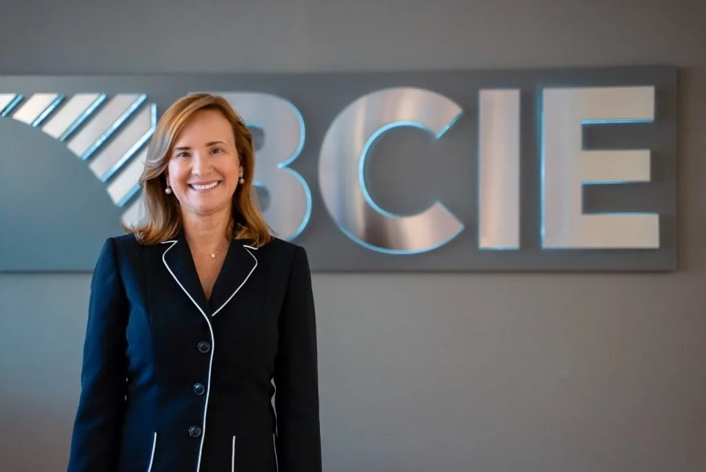 Mujer profesional sonriente vestida con traje formal oscuro frente al logotipo metálico de BCIE en una oficina.