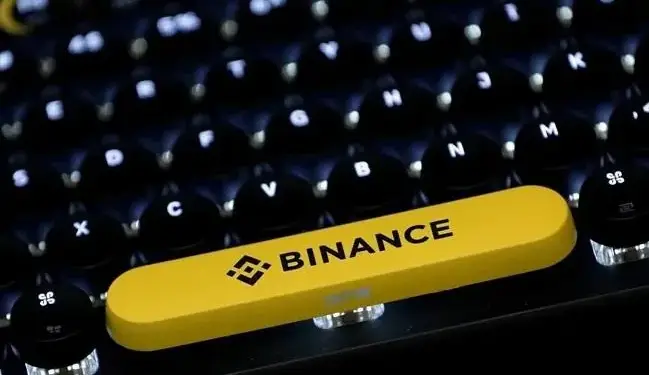Primer plano de un teclado de computadora con teclas negras retroiluminadas y una tecla rectangular amarilla con el logo de Binance y el texto 'BINANCE' en negro.