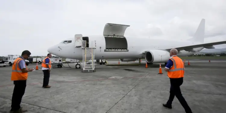 Avión de carga blanco con puertas abiertas en la pista, rodeado de personal de tierra con chalecos naranjas y equipos de apoyo. Muestra logística aérea.