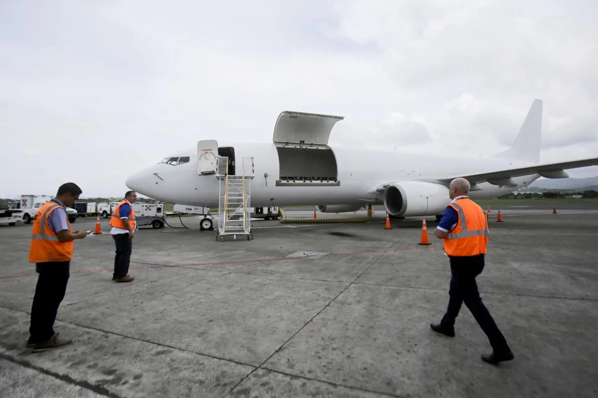 Avión de carga blanco con puertas abiertas en la pista, rodeado de personal de tierra con chalecos naranjas y equipos de apoyo. Muestra logística aérea.