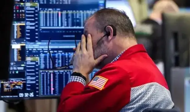 Operador financiero con chaqueta roja y bandera estadounidense cubriéndose la cara frente a múltiples pantallas con gráficos bursátiles.