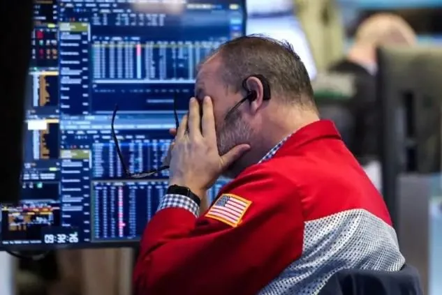 Operador financiero con chaqueta roja y bandera estadounidense cubriéndose la cara frente a múltiples pantallas con gráficos bursátiles.