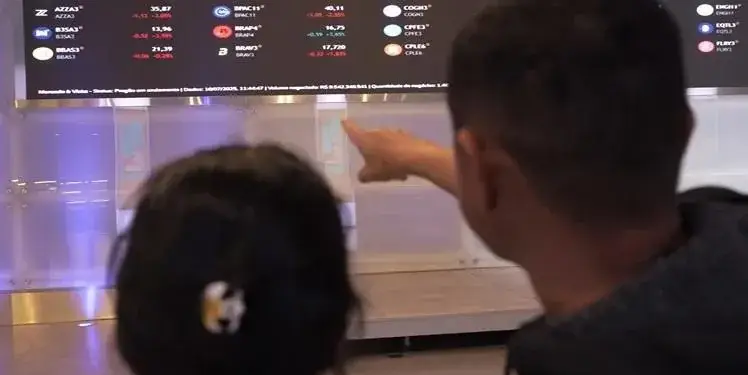 Dos personas observan una pantalla grande con datos del mercado de valores IBOVESPA B3, mostrando cotizaciones de acciones y sus variaciones. El hombre señala la información.