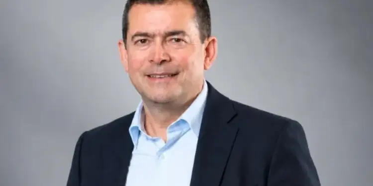 Retrato de un hombre de mediana edad, caucásico, con cabello oscuro y corto, sonriendo. Viste una chaqueta de traje oscura y camisa azul claro. Fondo gris liso.