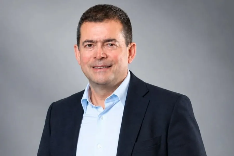 Retrato de un hombre de mediana edad, caucásico, con cabello oscuro y corto, sonriendo. Viste una chaqueta de traje oscura y camisa azul claro. Fondo gris liso.