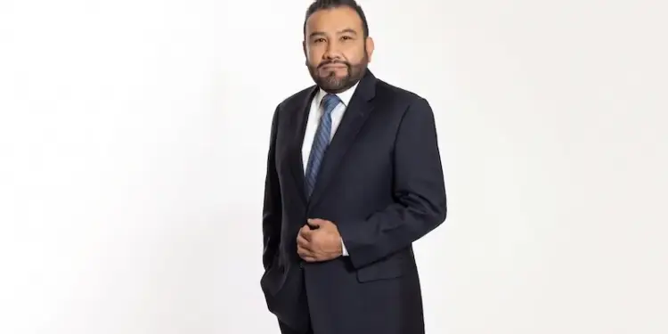 Hombre de negocios con traje oscuro, camisa blanca y corbata azul posando sobre un fondo blanco uniforme.