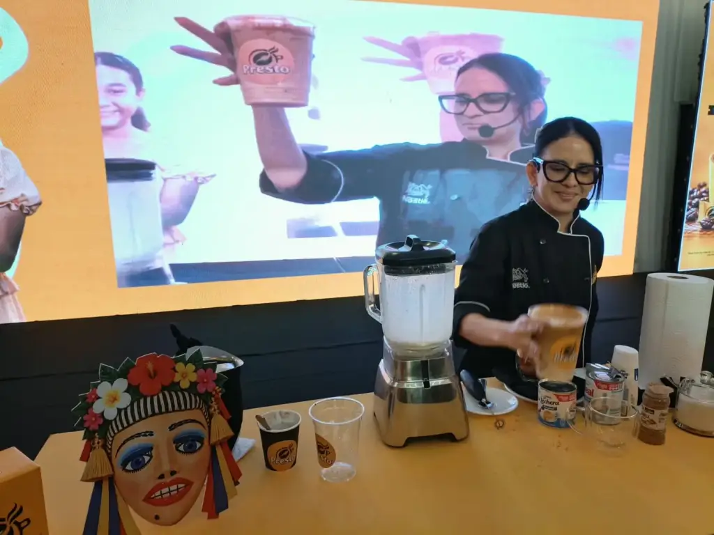 Una chef levanta una taza de melcochita preparada con Café Presto mientras la pantalla de fondo amplía la escena