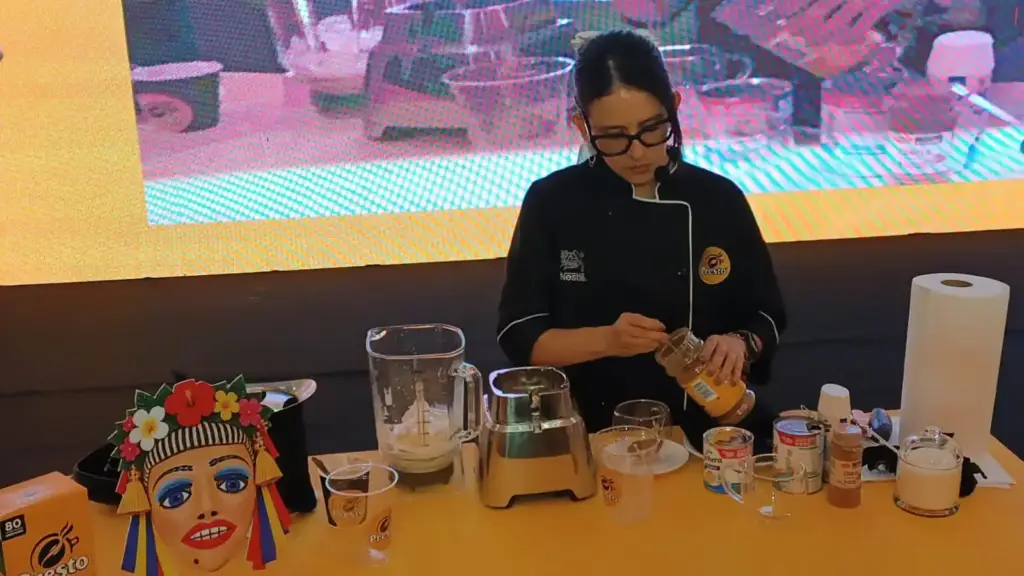 Una chef prepara café en una mesa con licuadora, tazas y frascos de Café Presto junto a una máscara del folclore nicaragüense