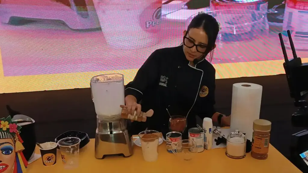 Una chef agrega ingredientes a una licuadora en una mesa con tazas y frascos de Café Presto