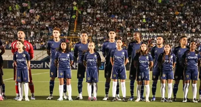 Jugadores de futbol con uniforme azul oscuro y niños acompañantes alineados en el campo antes de un partido con estadio lleno de espectadores.