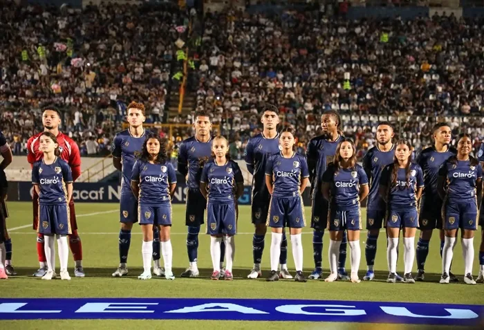 Jugadores de futbol con uniforme azul oscuro y niños acompañantes alineados en el campo antes de un partido con estadio lleno de espectadores.