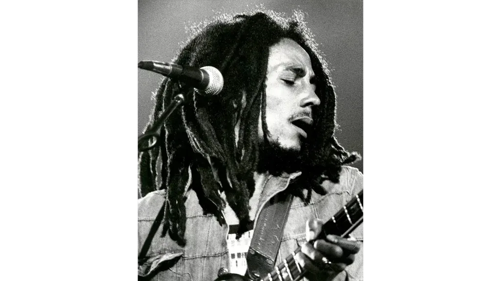 Fotografia en blanco y negro de un musico con rastas cantando frente al microfono y tocando la guitarra.