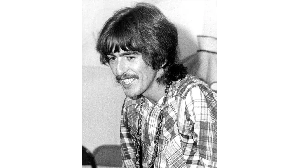 Fotografía en blanco y negro de un hombre sonriente con cabello largo, bigote y camisa a cuadros.
