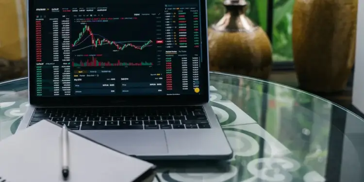 Laptop MacBook Pro mostrando gráficos de trading de criptomonedas con velas japonesas y datos de mercado. Un cuaderno y bolígrafo sobre mesa de cristal.