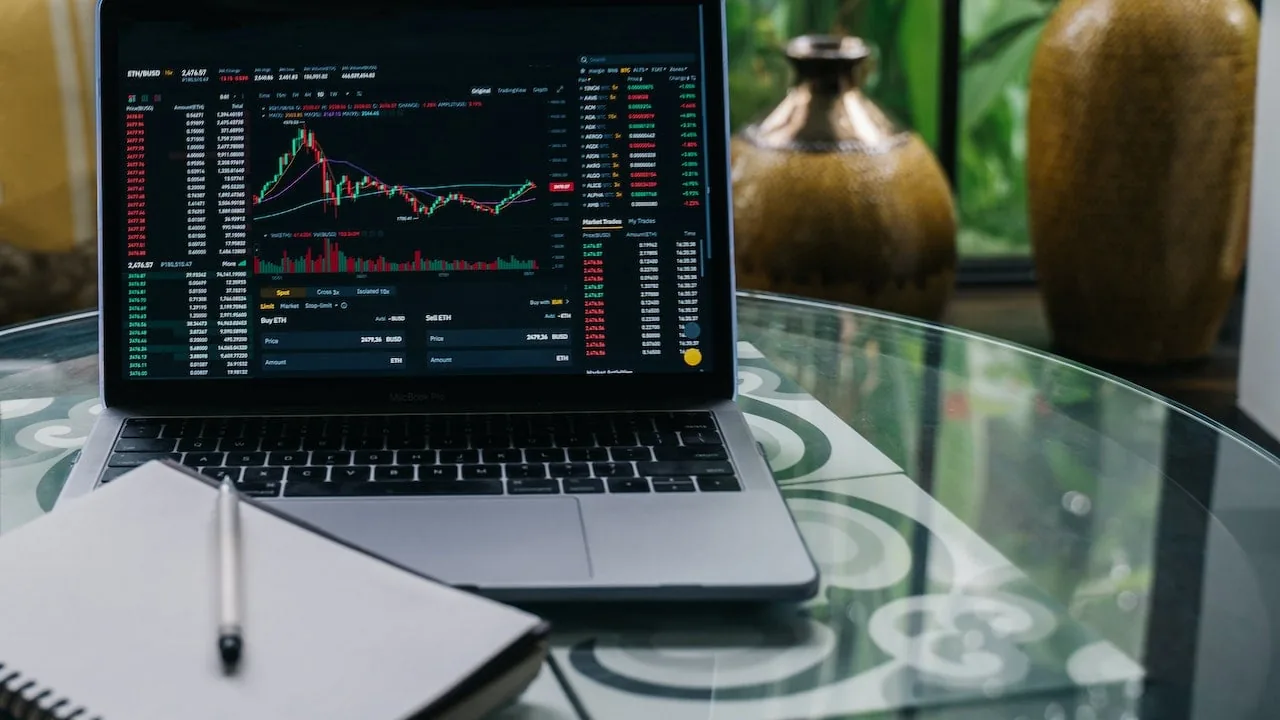 Laptop MacBook Pro mostrando gráficos de trading de criptomonedas con velas japonesas y datos de mercado. Un cuaderno y bolígrafo sobre mesa de cristal.